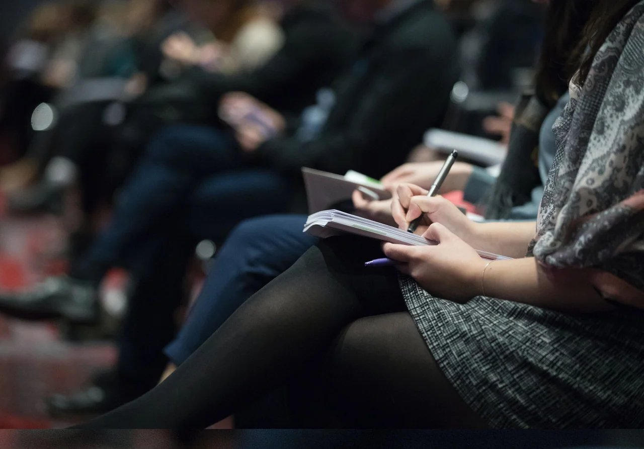 An audience seated in rows at an event, one person taking notes in a notebook