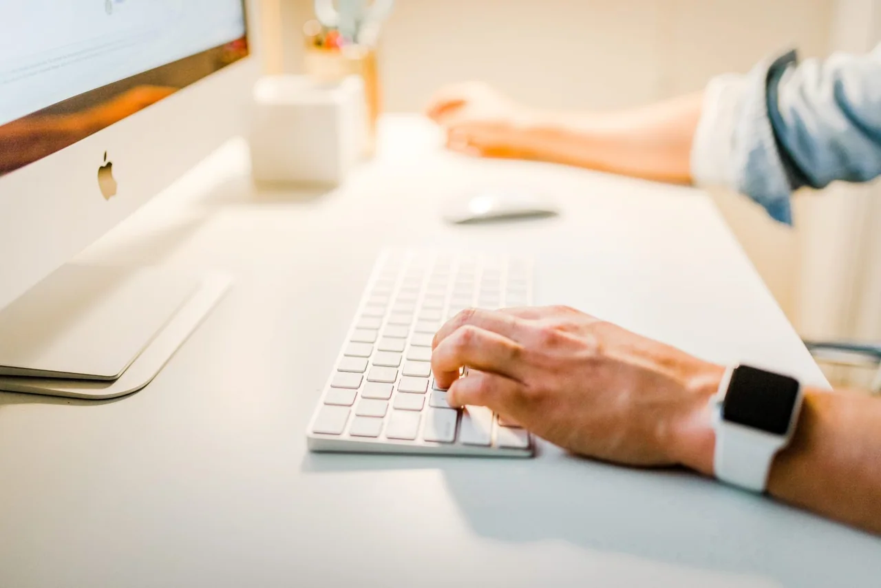 A hand typing on a keyboard in front of an iMac, on a white desk