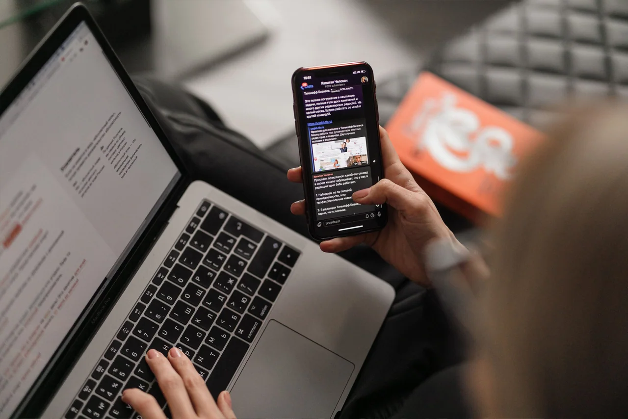 Someone using a laptop with one hand while checking a messaging app on their phone with the other