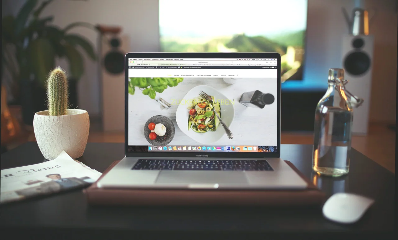 A laptop on a desk showing a food website with a salad on the homepage, next to a cactus, a newspaper and a glass bottle