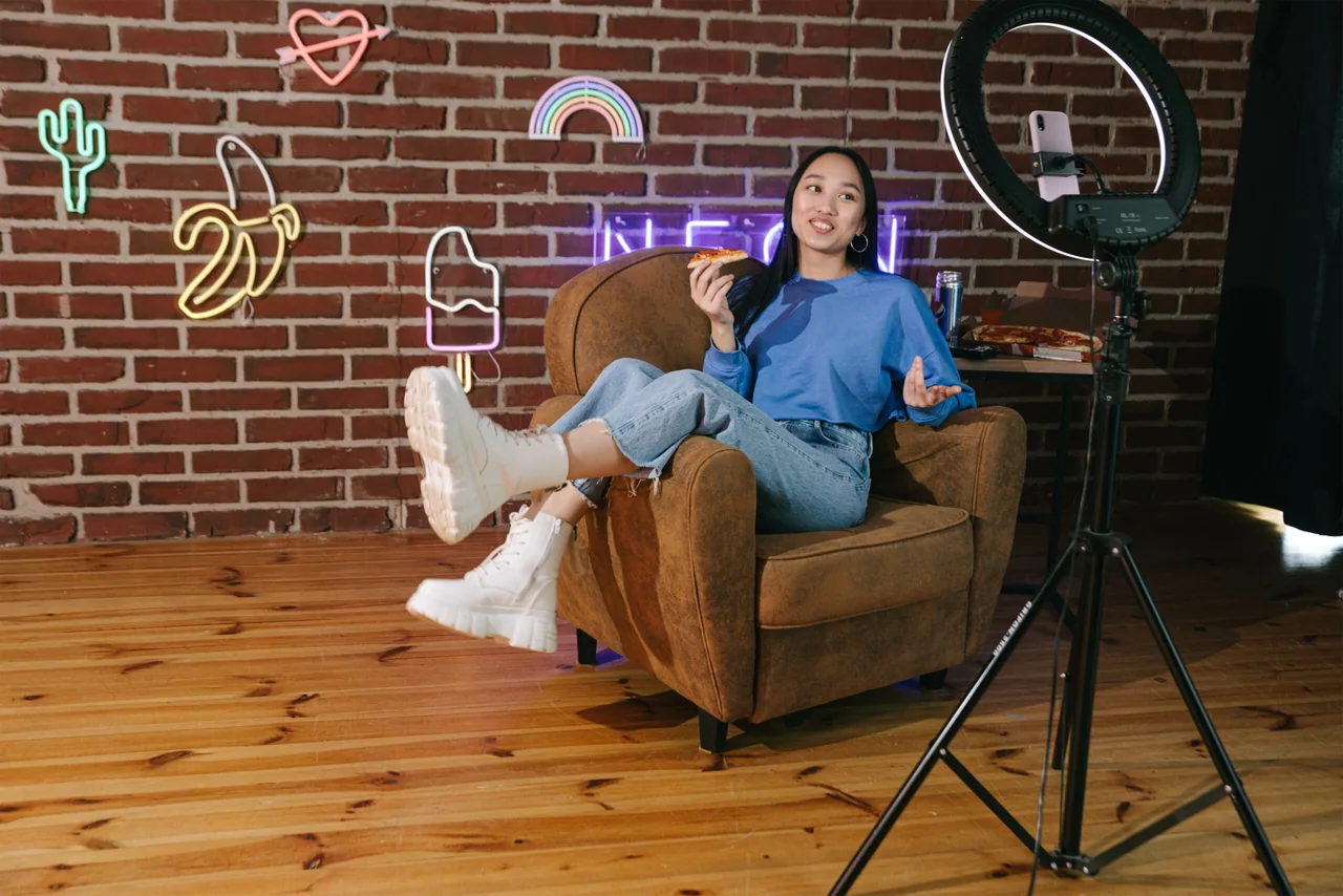A content creator filming herself in an armchair with a smartphone on a ring light and tripod, a neon-lit brick wall behind her