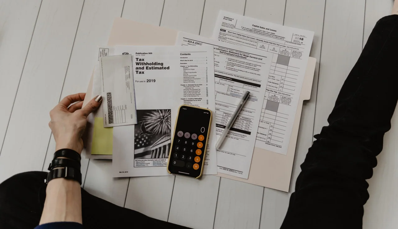 Tax forms, a calculator, a pen and an envelope spread across a desk, with a phone showing a calculator app