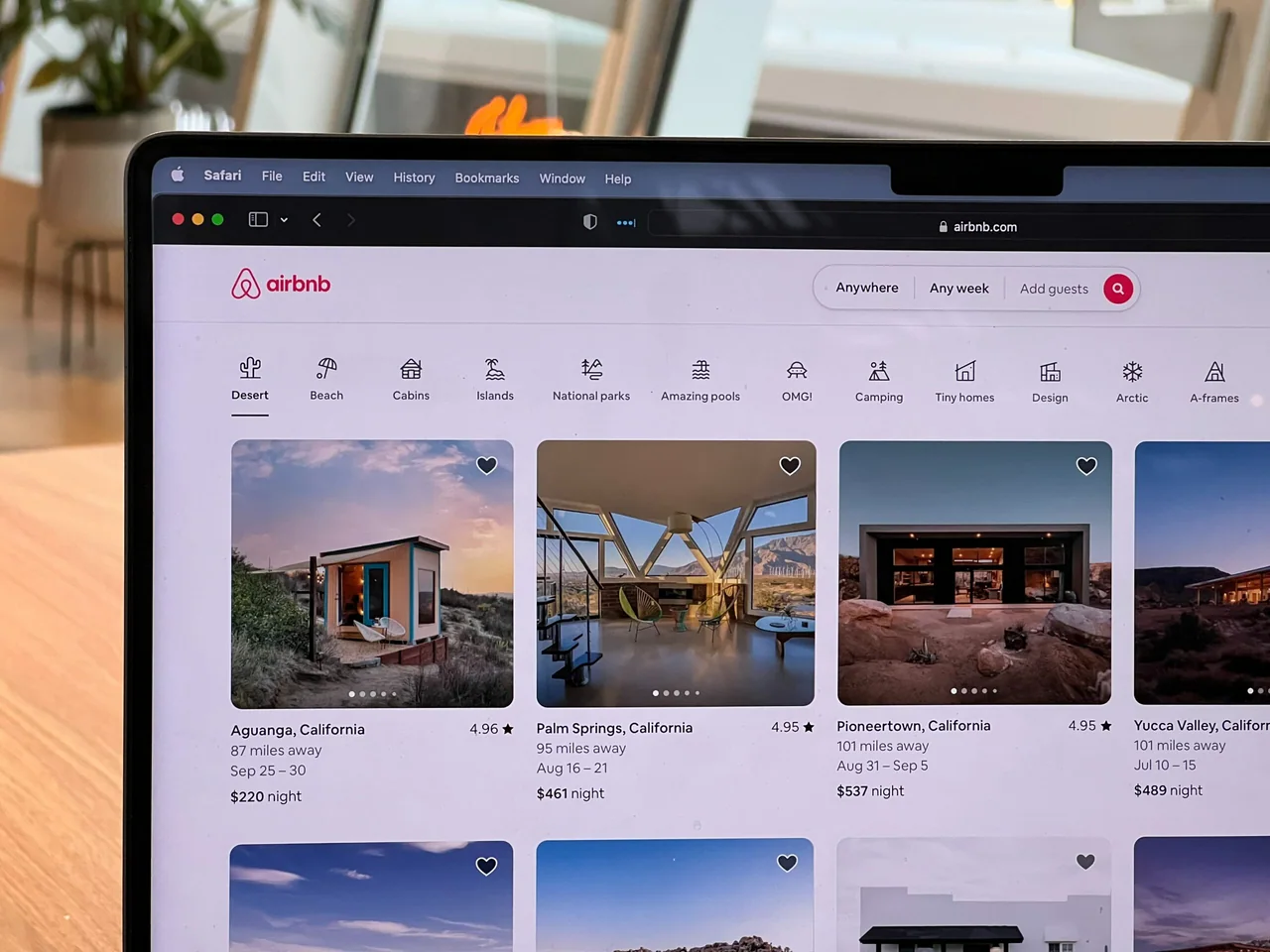 Laptop showing the Airbnb website with a grid of rental listings, photos, prices and star ratings