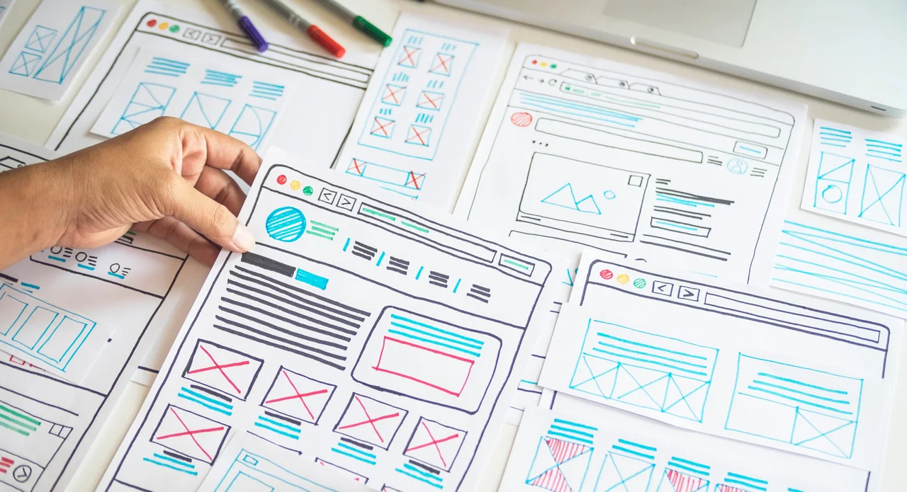 Hand-drawn wireframe sketches for website layouts spread across a desk with marker pens