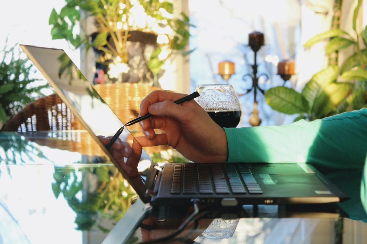 A person using a stylus on a laptop touchscreen at a table surrounded by plants