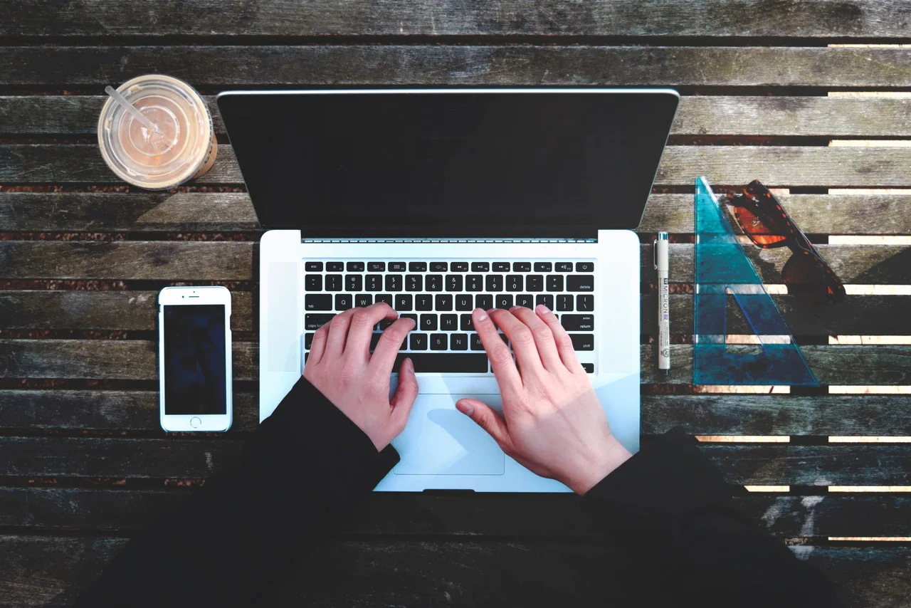 Hands typing on a laptop on a wooden table next to a phone and a drink